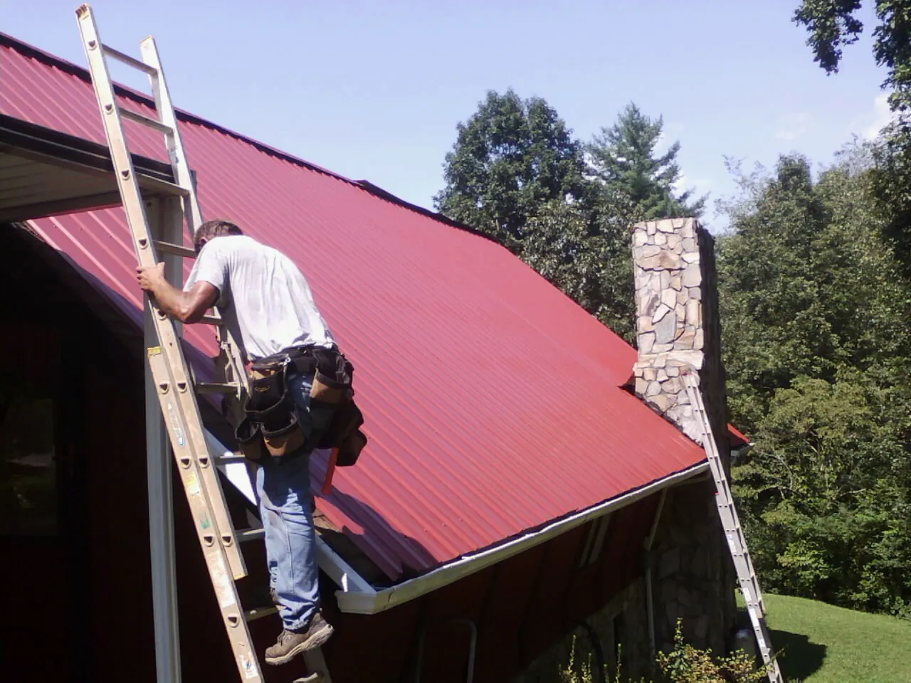 Roofer on a ladder beside a red metal roof and stone chimney — Soffit & Fascia Repair by Roofing Brothers in Sandisfield